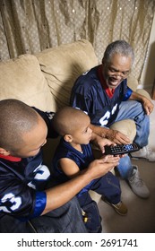 Three Male Generations Of An African-American Family Watching Football Game On Tv With Boy Holding Remote.