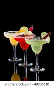 Three Margaritas - Apple, Orange And Raspberry - In Chilled Glasses Over Black Background, Garnished With Slice Of Green Apple, Limes, Orange Twist, Raspberry And Cinnamon Stick.