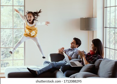 Three Members Of Diverse Family, Caucasian Father, Asian Mother And Little Half Daughter Spend Time Together In House Living Room. Girl Jumping On Sofa While Mom And Dad Working At Home And Cheer Up.