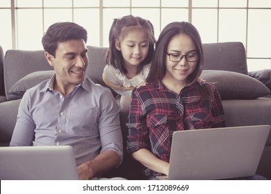 Drei Familienmitglieder, kaukasischer Vater und asiatische Mutter und kleine halbe Tochter sitzen zusammen im Wohnzimmer und benutzen Laptop-Computer. Idee für die Arbeit zu Hause.