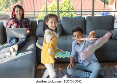 Three Members Of Family, Father, Mother And Kid Daughter Live Together In Home Living Room. Girl Throwing Toy With Dad While Mom Working With Notebook Computer. Idea For Work At Home And Life Balance.