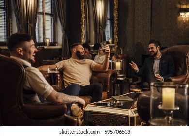 Three Men Are Enjoying Drinks In A Bar Lounge. They Are Talking And Laughing While Drinking Pints Of Beer. 