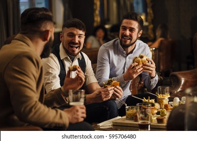 Three men are sitting together in a bar/restaurant lounge. They are laughing and talking while enjoying burgers and beer. 