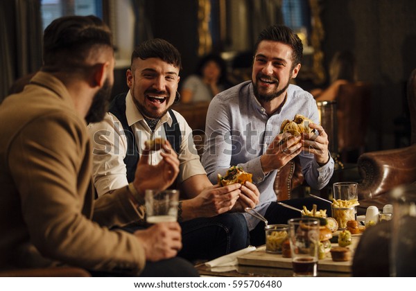 Three men are sitting together in a bar/restaurant
lounge. They are laughing and talking while enjoying burgers and
beer. 