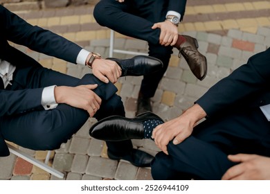 Three men in suits are sitting on a brick sidewalk, each holding the other's shoe. The men are wearing black dress shoes and are wearing ties. The scene is casual and relaxed - Powered by Shutterstock