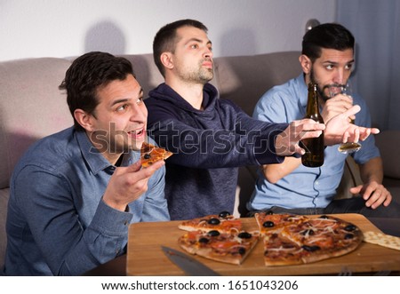 Three men watching matchup on tv with anxious uncertainty while drinking beer together at home