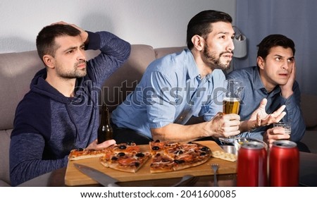 Three men watching matchup on tv with uncertainty serious while drinking beer together at home