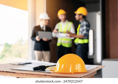 Three men wearing hard hats are standing around a table with blueprints and a yellow hard hat - Powered by Shutterstock