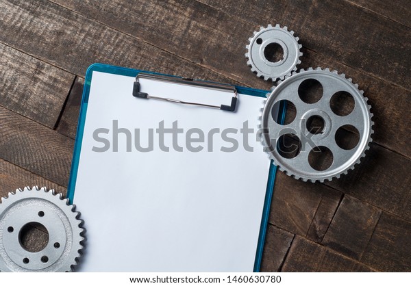 Three metal silver gears lies near blue plastic\
clipboard with empty paper sheets on old weathered wooden table in\
workshop. Copy space for\
text