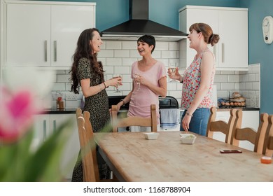 Three Mid Adult Women Standing In A Kicthen Enjoying A Glass Of Wine. The Women Are All Standing Around The Stove In The Kitchen, Talking And Laughing.