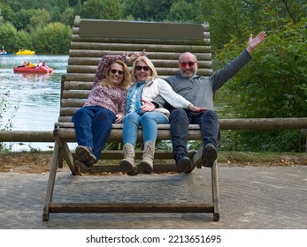 Three Middle Aged People Having Fun Times And Laughs Whilst Sat On A Giant Outdoor Seat. Laughing And Waving They Pose For A Holiday Photograph. Summer Vacation As Good Friends Celebrate Together.