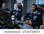 Three middle aged security guards including Black man and two Caucasian men monitoring surveillance screens in control room, sitting at desks with focused expressions, wearing uniforms and badges