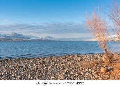 Three Mile Beach With View Of Okanagan Lake And Distant Snow Covered Mountains With Blue Sky