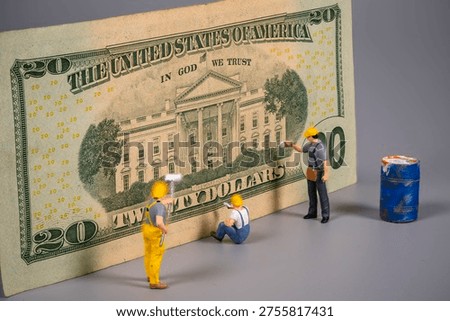 Three miniature construction workers are seen near a twenty dollar bill. One worker paints, another sits, and the third stands by a blue paint can. This scene shows work on money.