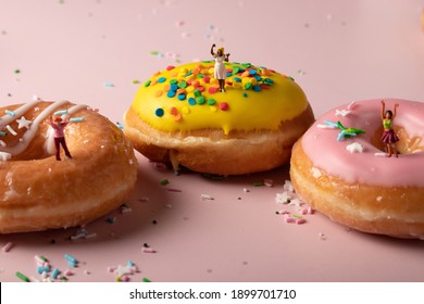 Three miniature women celebrating on glazed, colourful donuts. Donut party or coffee break at work. Binge eating or snacking on unhealthy sweets. The joy of a treat. Girls cheer on delicious pastries.