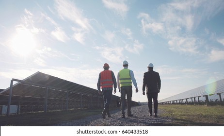 Three Mixed Ethnic Men In Uniform And Hard Hat Walking Around Solar Power Station And Examining It. Side View