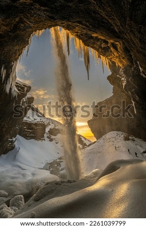 Three mountaineering tourists in front of the frozen Kvernufoss waterfall, with snow and stalagmites and falling water illuminated by the first rays of the sunrise or sunset sun reflecting its light