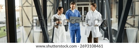 Three multicultural doctors engage in discussion while reviewing a laptop in a hospital corridor, banner