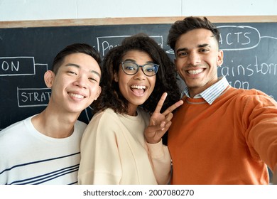 Three Multicultural Young Happy Cheerful Diverse College Students Gen Z Team Having Fun Together Making Selfie On Black Board In Classroom. International Diversity Concept. Phone Camera View