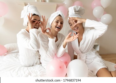 Three multi-ethnic beautiful well-groomed brides maids women wearing bathrobes with towels on head making binoculars with fingers celebrating bachelorette party room decorated with pink white balloons