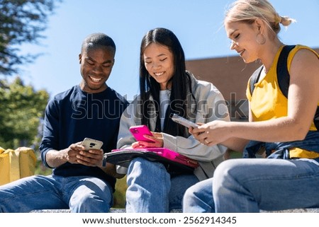 Three multiethnic students are using their smartphones while sitting on a wall on a sunny day on campus