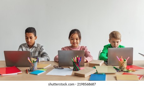 Three Multiracial School Kids Using Laptops Learning Online Sitting At Desk In Modern Classroom At School. Schoolboys And Schoolgirl Having Class Browsing Internet On Computers. Panorama