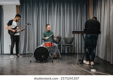 Three musicians practice in a studio, focusing on guitar, drums, and keyboard. They are setting up equipment and preparing for a session. The room is casual with curtains as the backdrop.