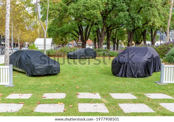 Three mystery cars draped under\
black covers in a park setting in Melbourne,\
Australia