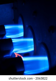 Three Natural Gas Burners Firing Brilliant Blue Flames Into An Active Furnace, The Core Of A Central Heating System. Orange Pilot Light/filament At Lower Left Ensures Burners Stay Lit.