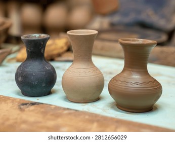 Three newly made small ceramic vases in different shades of clay stand in a row on a pottery studio workbench.