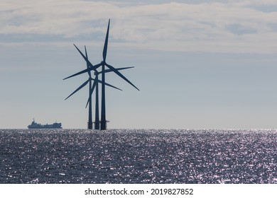 Three offshore wind farm wind turbines in patterned silhouette. Sea horizon with glistening water dominated by a dark geometric pattern of crossed wind turbine blades and a distant passing ship.