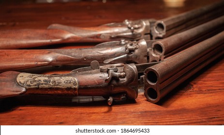 Three old, vintage, break-action double-barrel shotguns featuring two triggers and two hammers sit on a wooden table. Their bluing faded to a patina.