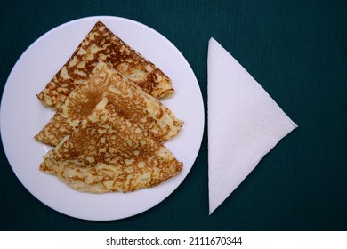 Three Pancakes Folded In A Triangle Lie On A White Plate. On The Right Is A White Napkin.