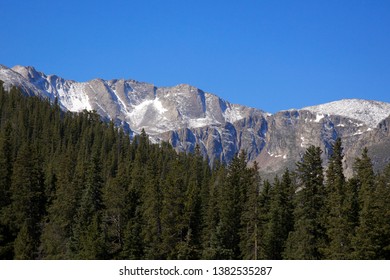 Three Part Scene Of A Clear Blue Sky Over A Rocky Snow Covered Mountain Over A Lush Dark Green Evergreen Treeline