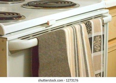 Three patterns of dish towels hang off an oven's handle.  Electric burners are in the upper portion of the image. Light brown cabinets to the right.