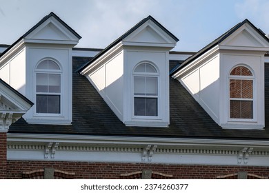 Three peaked dormer-style windows on a hip roof covered with black shingles. The vintage white wooden frames have arch-shaped glass and closed double-hung panes with white curtains hanging inside.  