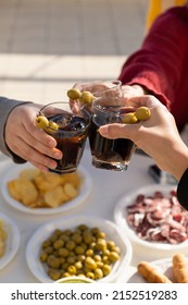 Three people clinking with vermouth glasses in a sunny day. Just three arms cropped above a table full of snacks. Friends enjoying drinks and food.