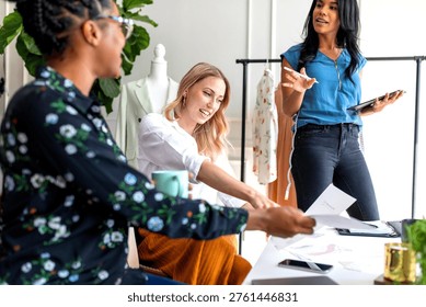 Three people in a creative office setting, discussing documents. A woman in a blue shirt stands, while others sit, surrounded by fashion elements.