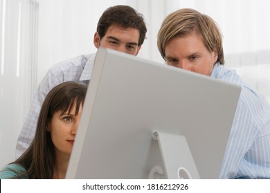 Three People Looking At A Computer Screen At An Office Desk.