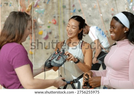 Three people are preparing for climbing inside a gym. Two women are discussing climbing shoes while a man climbs in the background.