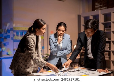 Three people are sitting at a table, looking at a piece of paper. They are all dressed in business attire, and one of them is holding a pen. Scene is serious and focused