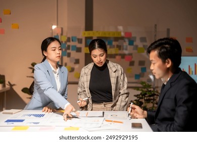 Three people are sitting at a table with a whiteboard in front of them. They are discussing something and one of them is pointing at the board. Scene is serious and focused