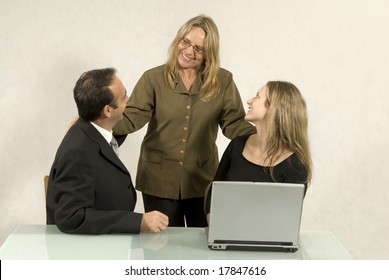 Three People Are Together In A Room At A Table. The Older Woman Is Standing And The Other Two Are Sitting Down.