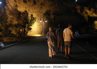 Three People Walking Away Along A Marina In The Amalfi Coast, Focused On The People And Blurring The Distant Cliff Face