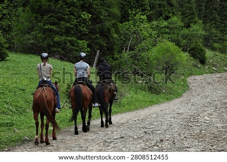 Three people wearing protective helmets ride brown horses away from the camera along a rugged gravel pathway. The outdoor scene captures adventure tourism, equestrian recreation, and active lifestyle in a scenic mountain valley.
