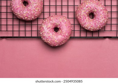 Three pink frosted donuts with multicolored sprinkles rest partially on a cooling rack, set against a pink background, exuding an inviting and delicious appearance. - Powered by Shutterstock