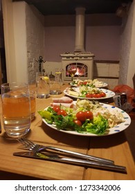 Three Plates Of Healthy Food And Glasses Of Water Lined Up On A Wooden Table In Front Of A Lit Stove Fire.