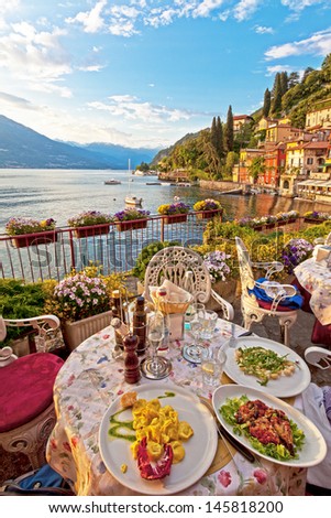 Three plates of lovely vegetarian Italian food on white plates on a table on a lovely outdoor terrace overlooking a beautiful, calm lake, with antique buildings and alpine mountains in the background