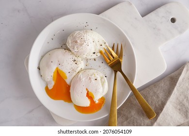Three poached eggs with egg yolk on a white plate on a marble board and golden forks
