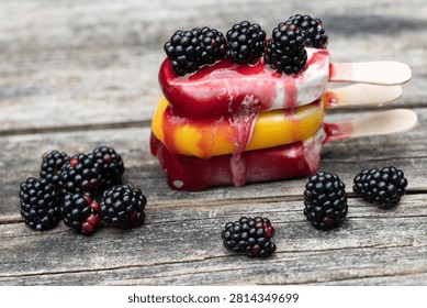 Three popsicles are stacked on top of each other on a rustic wooden board. The popsicles are starting to melt. There are blackberries on top of and next to the popsicles.